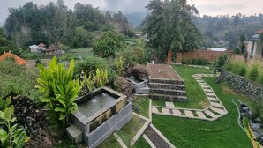 Un jardin luxuriant agrémenté d’une fontaine en pierre, d’un sentier sinueux pavé de pierres et offrant, en arrière-plan, une vue sur un paysage vallonné.