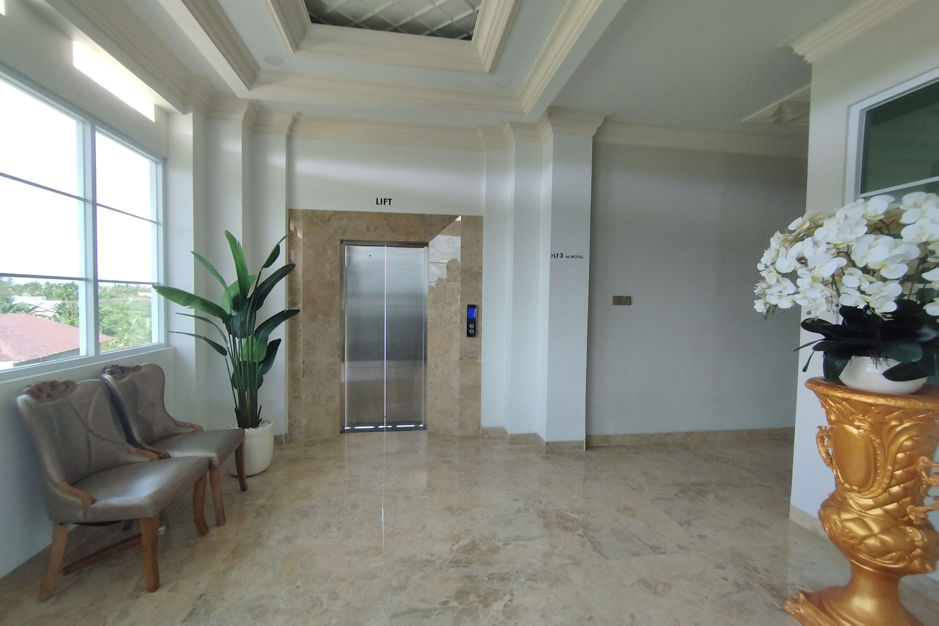 A spacious lobby with a marble floor, a lift, two wooden chairs, a potted plant, and a decorative golden vase.