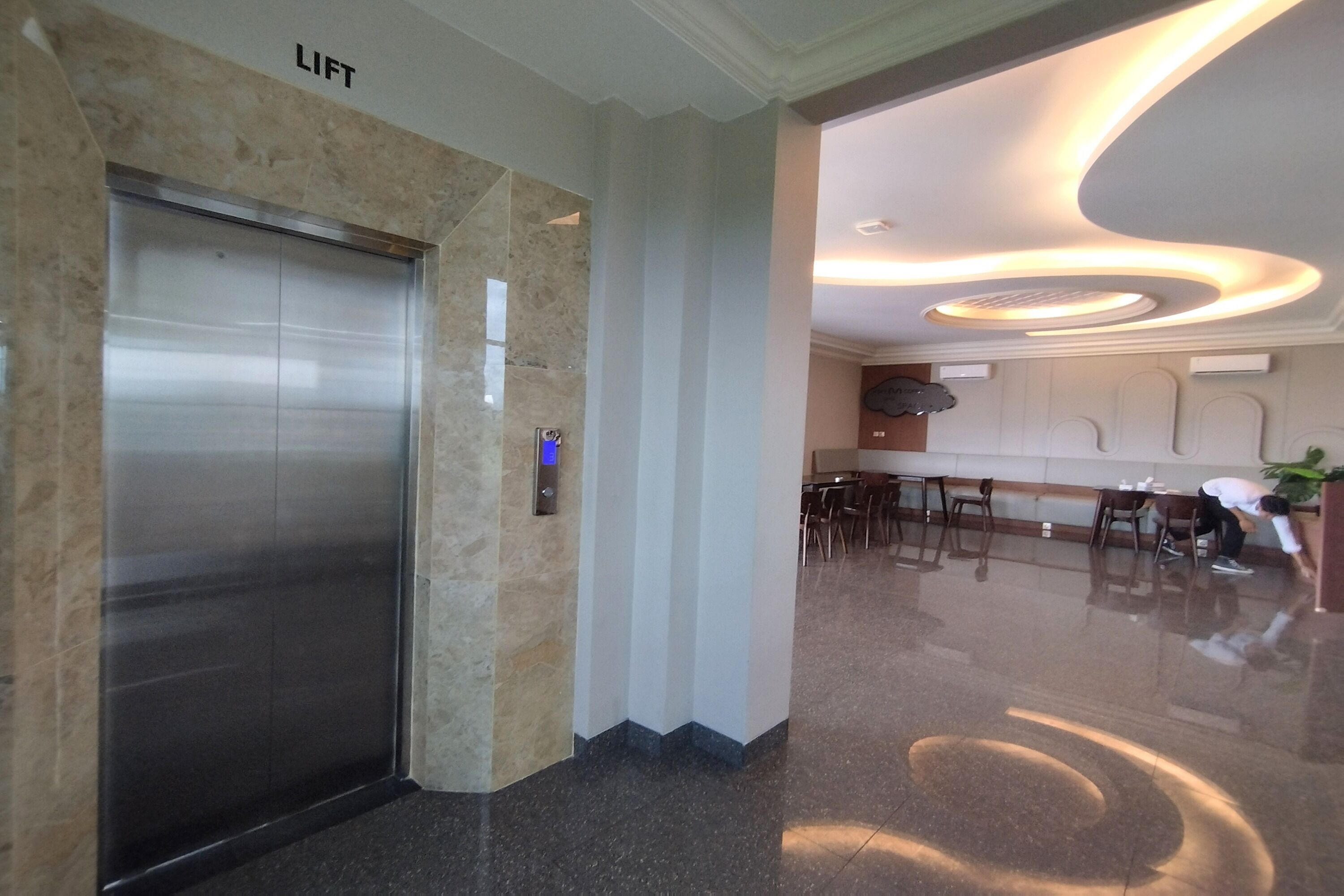 A modern elevator with a 'LIFT' sign, a well-lit reception area with a curved ceiling, and a seating area with tables and chairs.