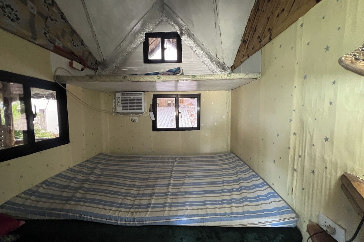 A small, single-room accommodation with a sloped ceiling, two windows, and a striped mattress.