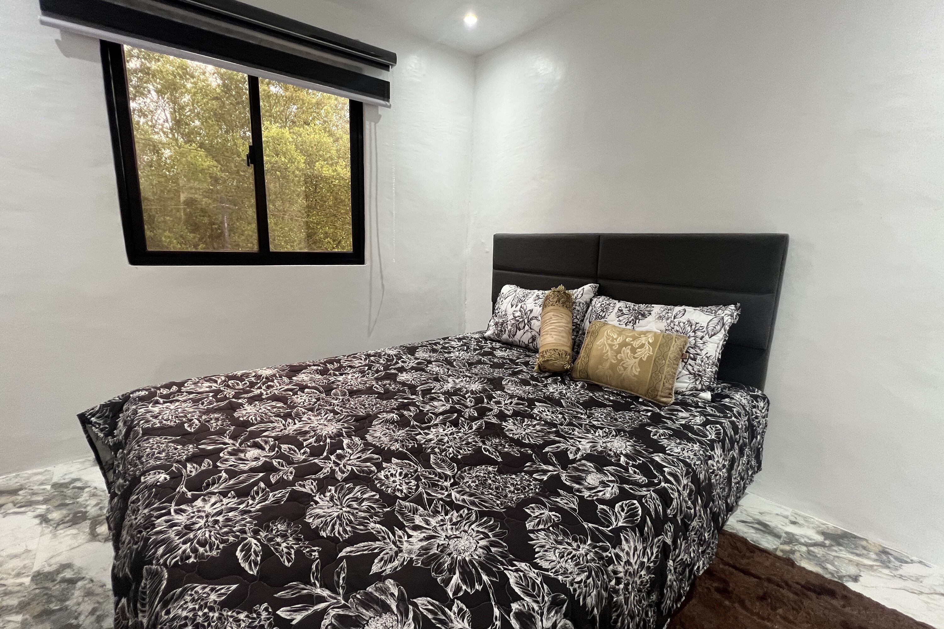 A bedroom with a black and white floral bedspread, a dark headboard, and a window with a view of trees.