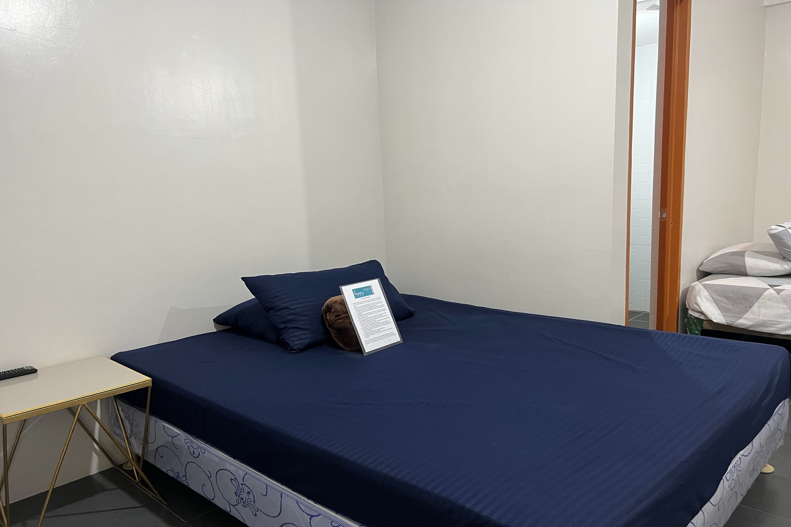Una cama individual con una colcha azul oscuro y un almohadón, una mesita de noche pequeña, y un cartel sobre la cama.