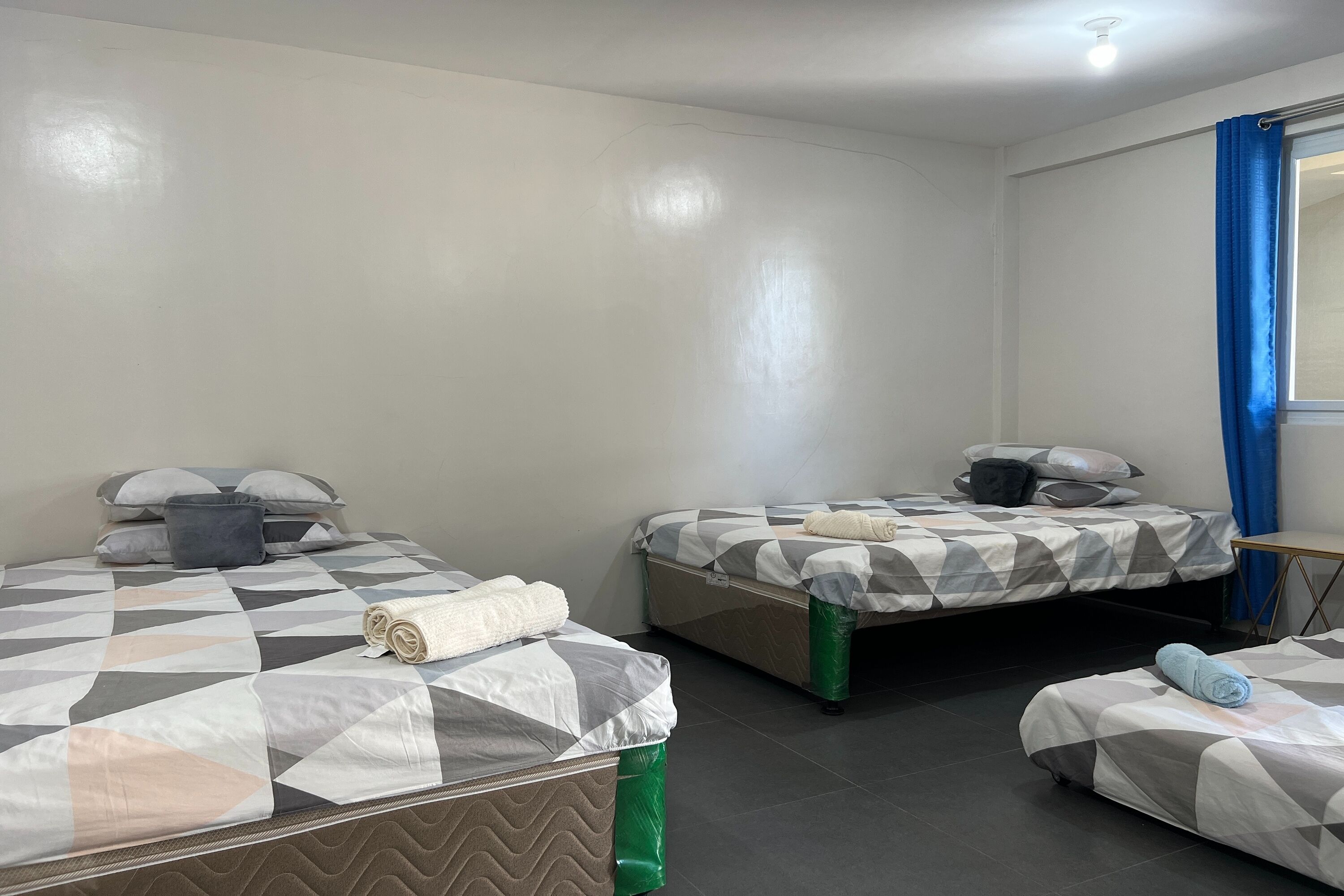 Ein Hotelzimmer mit zwei Betten, jeweils mit einem gemusterten Bettbezug, einem kleinen Tisch und einem Fenster mit blauen Vorhängen.