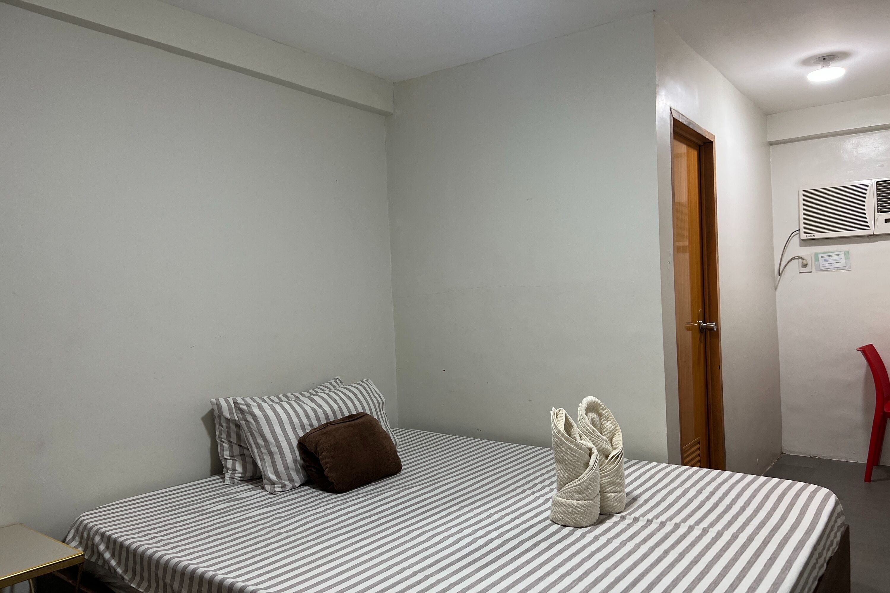 A single-bed room with a striped bedspread, a brown pillow, and a towel on the bed. There is a wooden door, a red chair, and an air conditioning unit on the wall.