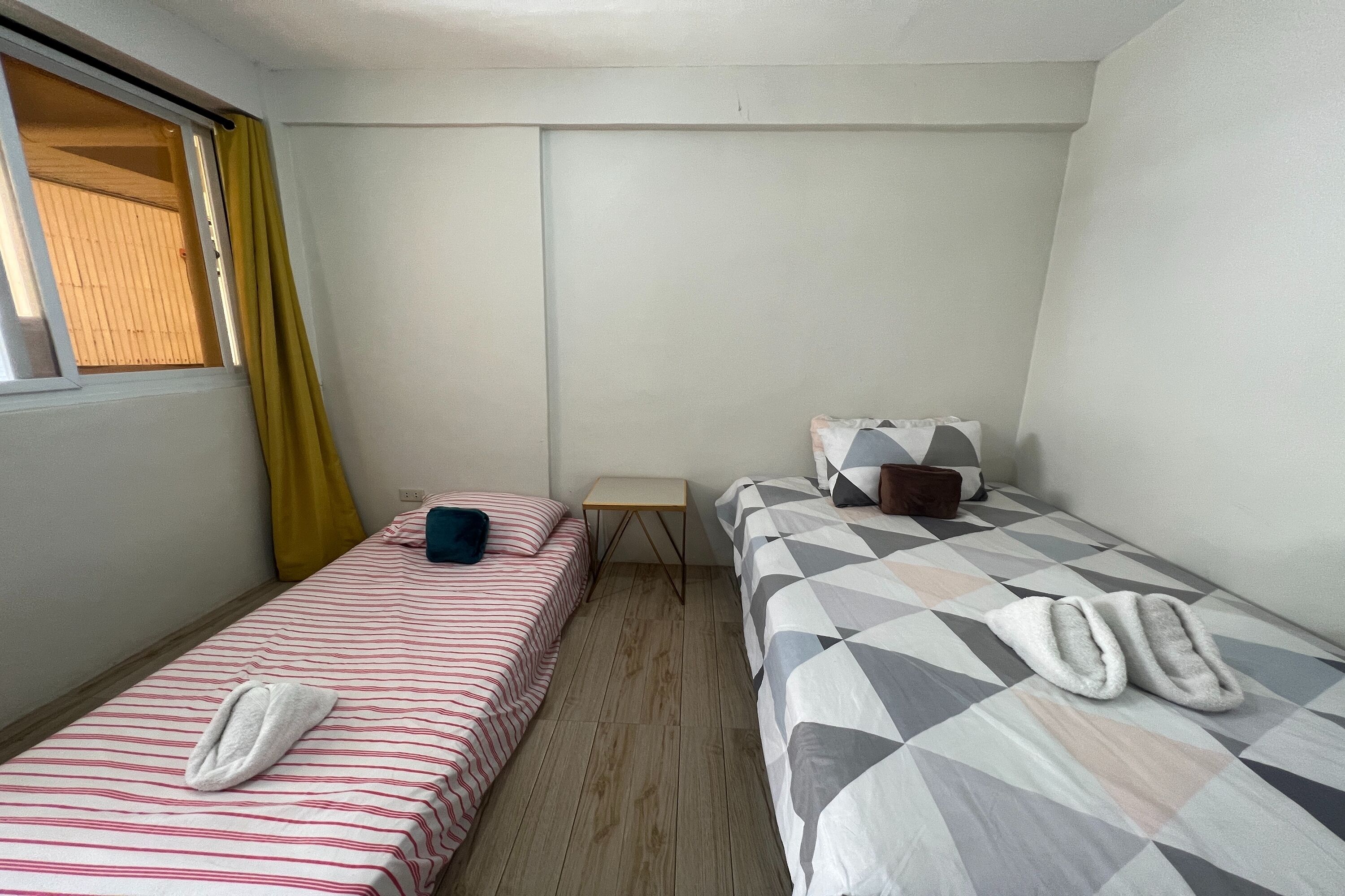 A small, clean room with two beds, a small table, and a window with yellow curtains.
