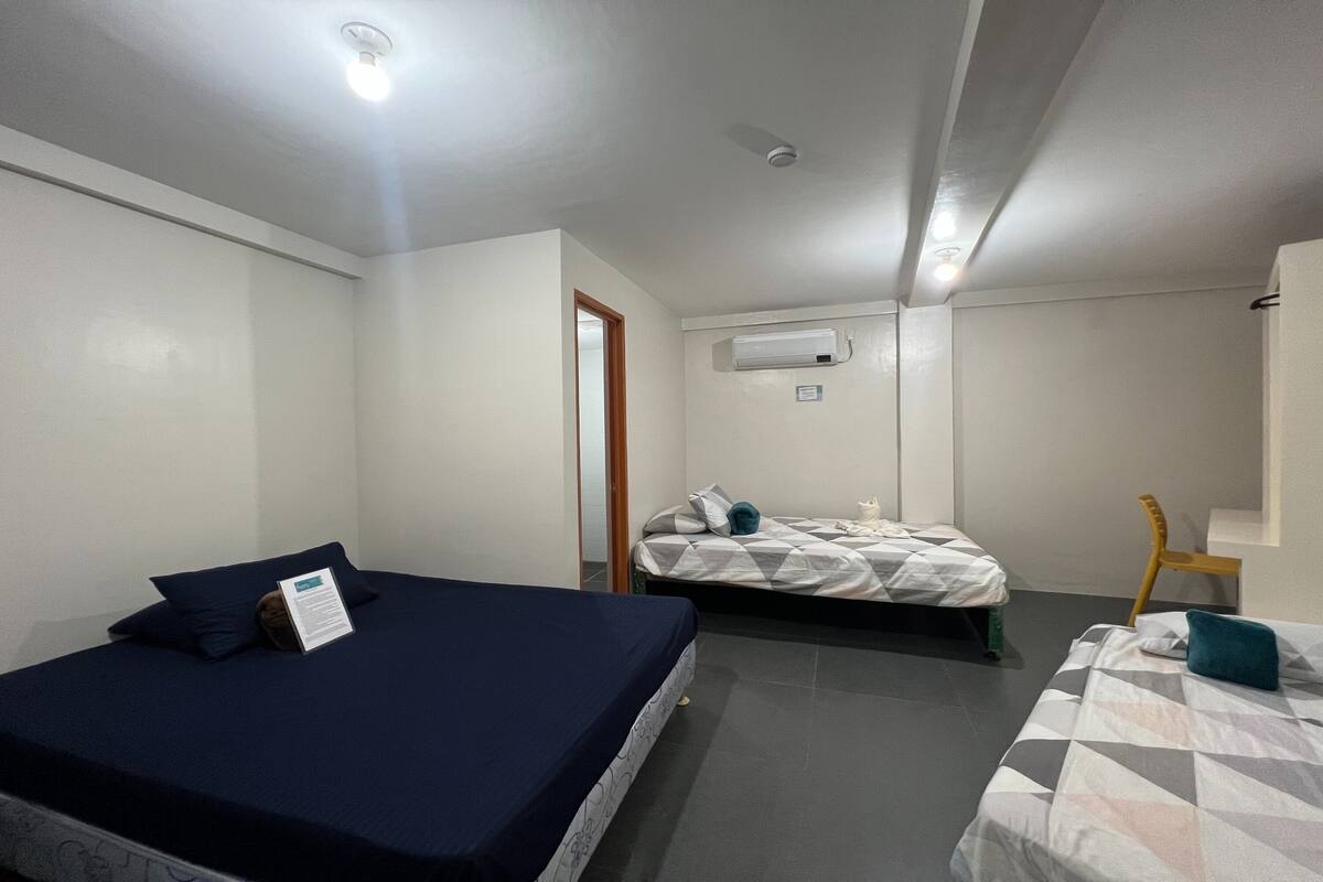 A small, clean room with two beds, a wooden chair, and a white air conditioning unit on the wall.