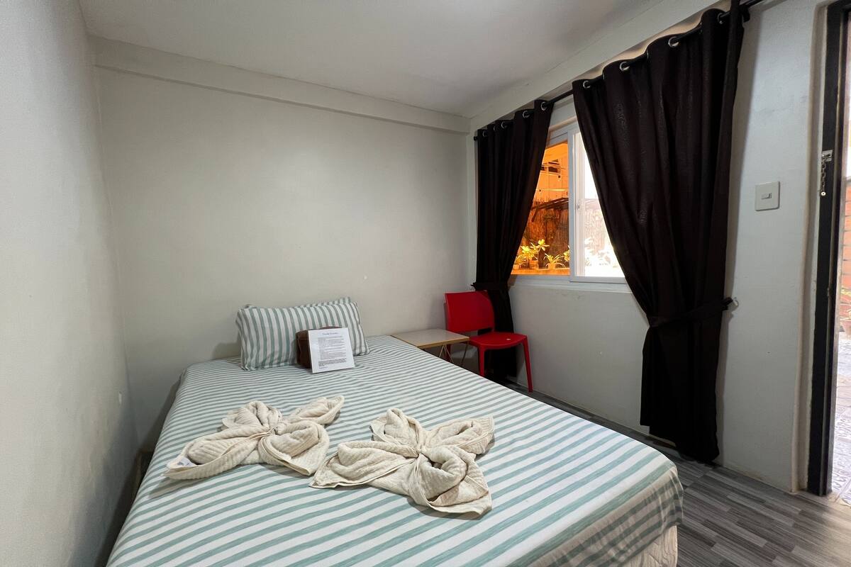 A hotel room with a bed, a red chair, a window with curtains, and a view of the outside.