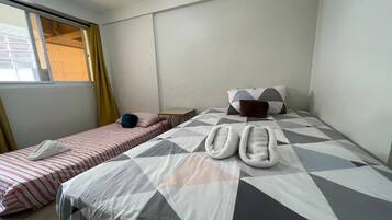 Une chambre d’hôtel avec deux lits, l’un recouvert d’une couvre-lit à rayures et l’autre d’une couvre-lit à motifs géométriques. On trouve des chaussons sur le lit à motifs géométriques, une petite table et une chaise.