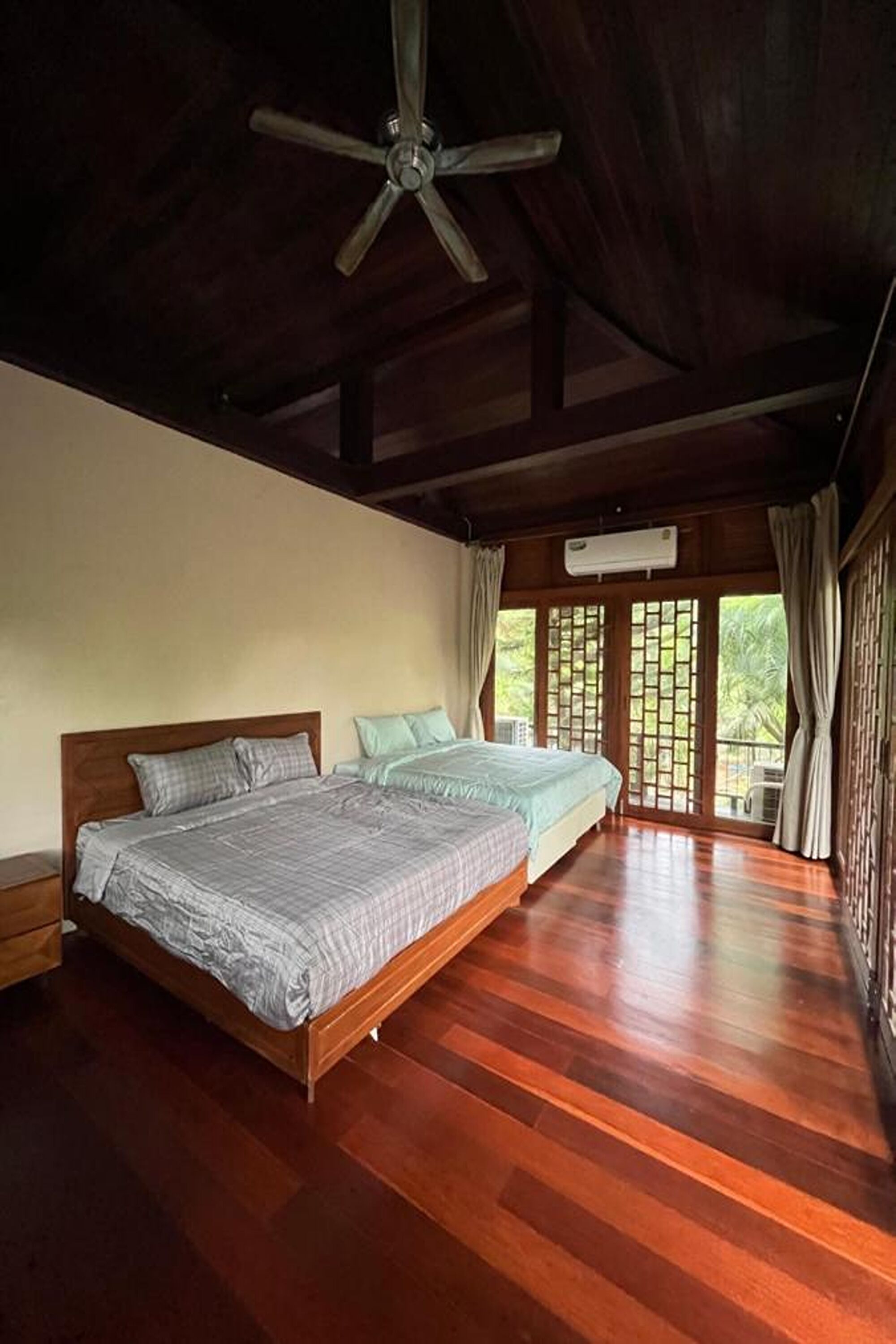 A spacious bedroom with two beds, wooden flooring, a ceiling fan, and large windows with curtains.