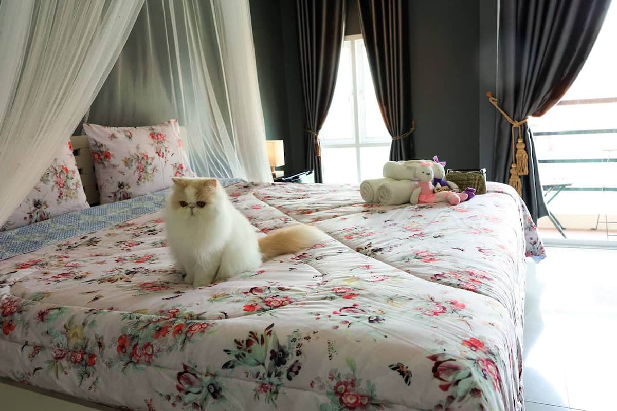 A bed with a floral bedspread, a cat sitting on it, and a teddy bear beside the bed.