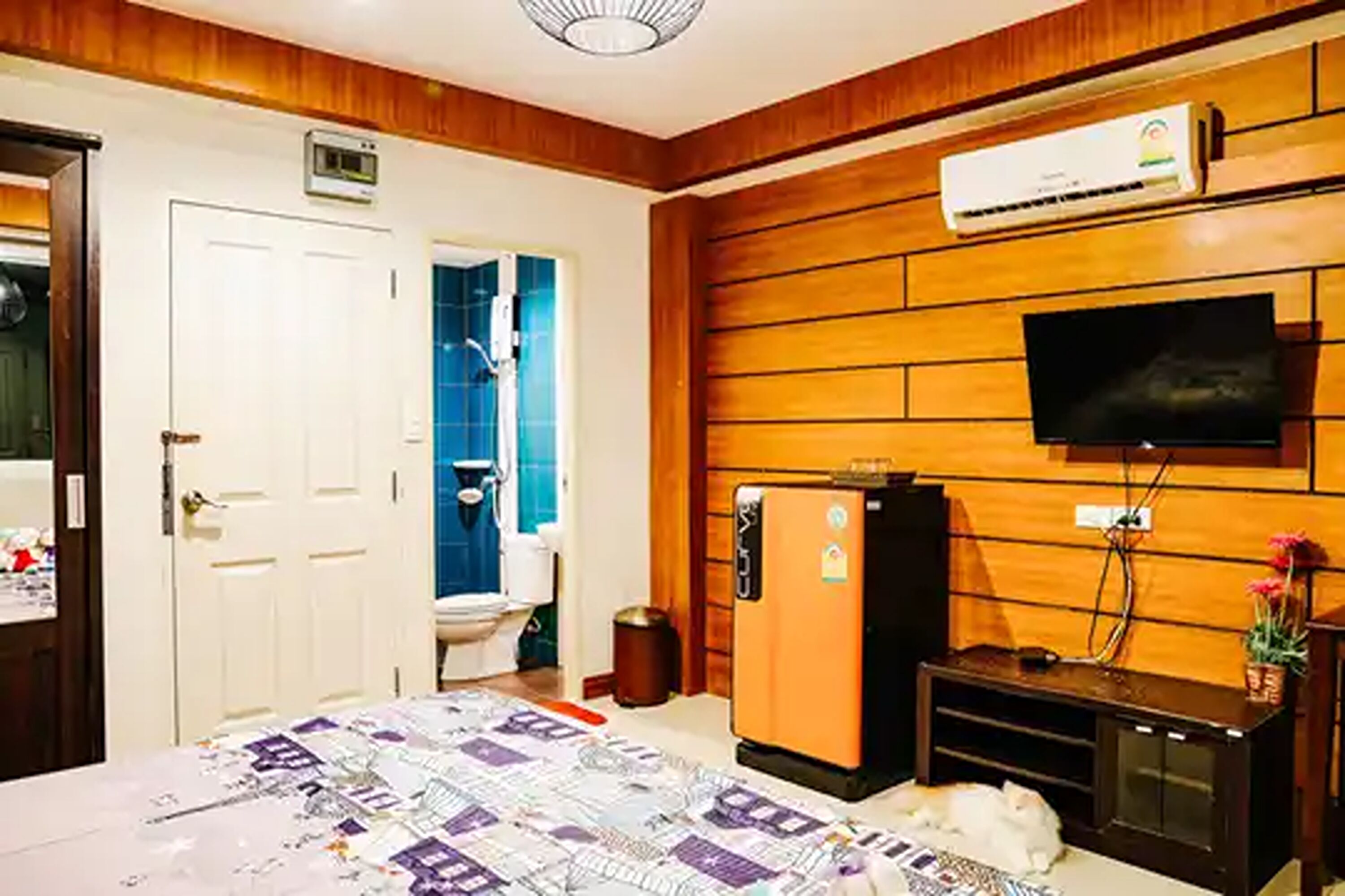 Quarto de hotel com lambris de madeira, uma cama com colcha estampada, uma televisão, um frigorífico e uma casa de banho com azulejos azuis.
