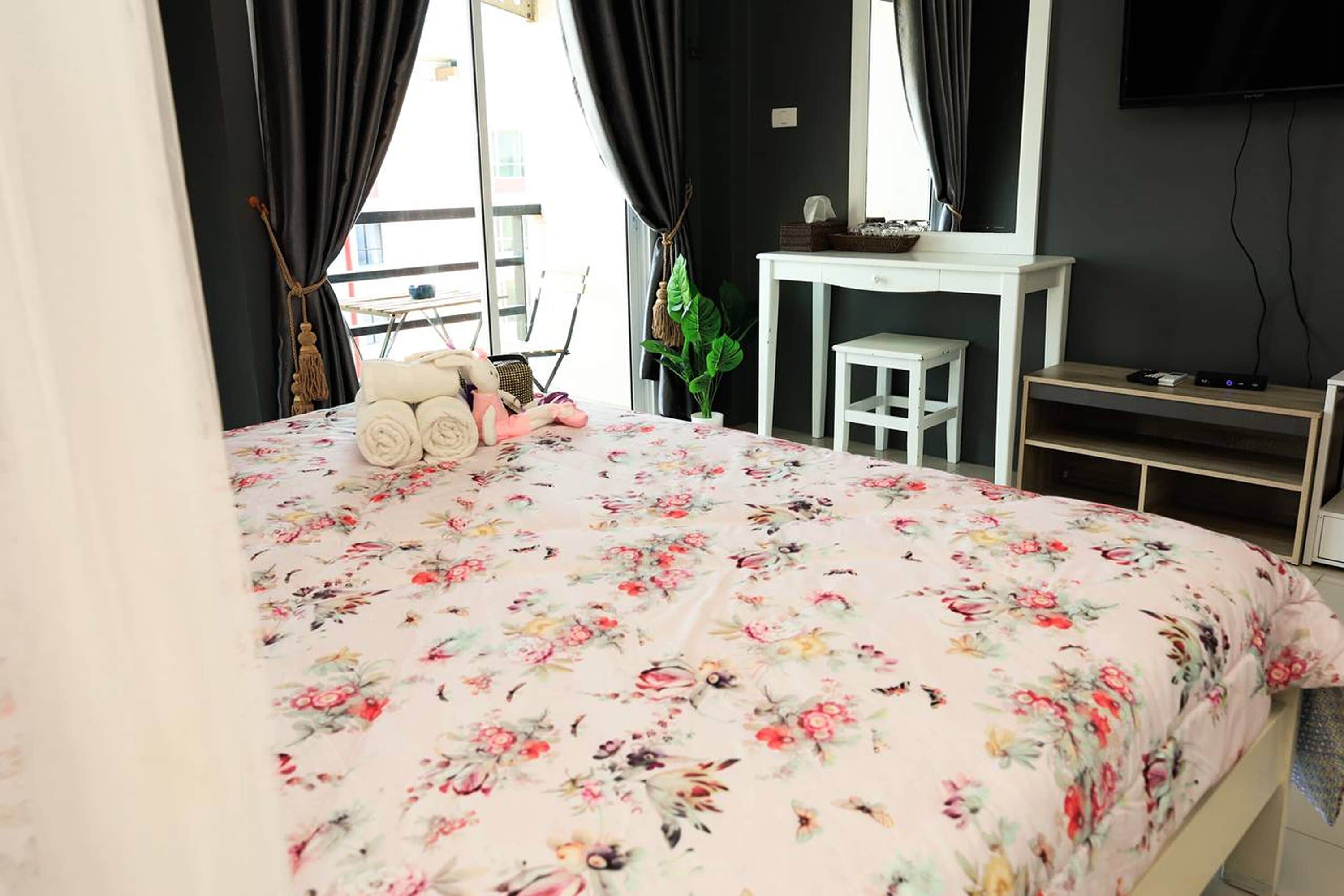 Ein Schlafzimmer mit Blumenmuster-Bettdecke, einem weißen Nachttisch, einem Fernseher und einem Fenster mit schwarzen Vorhängen.