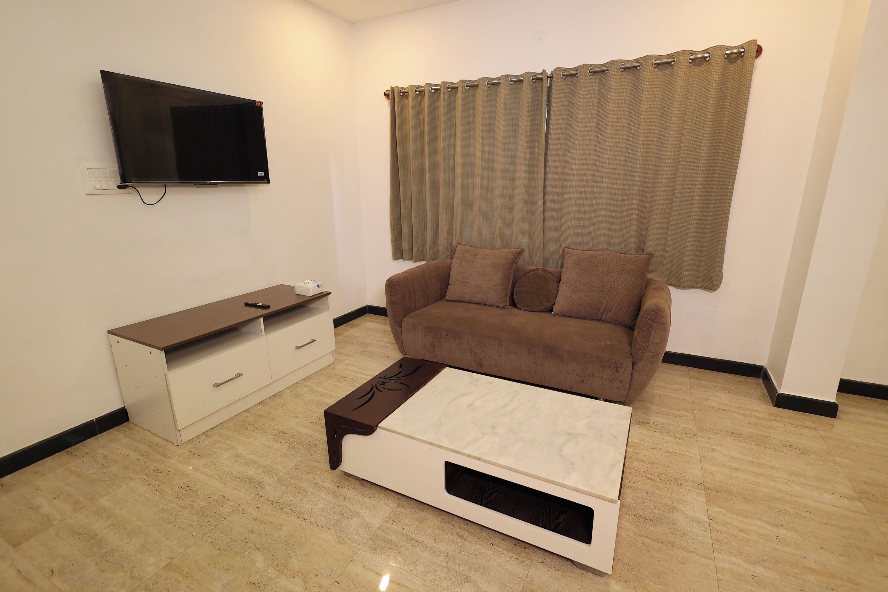 A living room with a flat-screen TV mounted on the wall, a brown sofa with cushions, a white coffee table with a drawer, and a window with beige curtains.