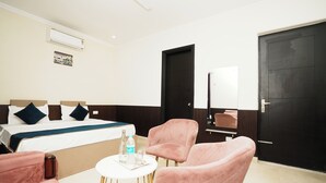 Habitación de hotel con una cama, dos sillas rosas, una mesita redonda con una botella y vasos, una puerta negra y un equipo de aire acondicionado blanco en la pared.