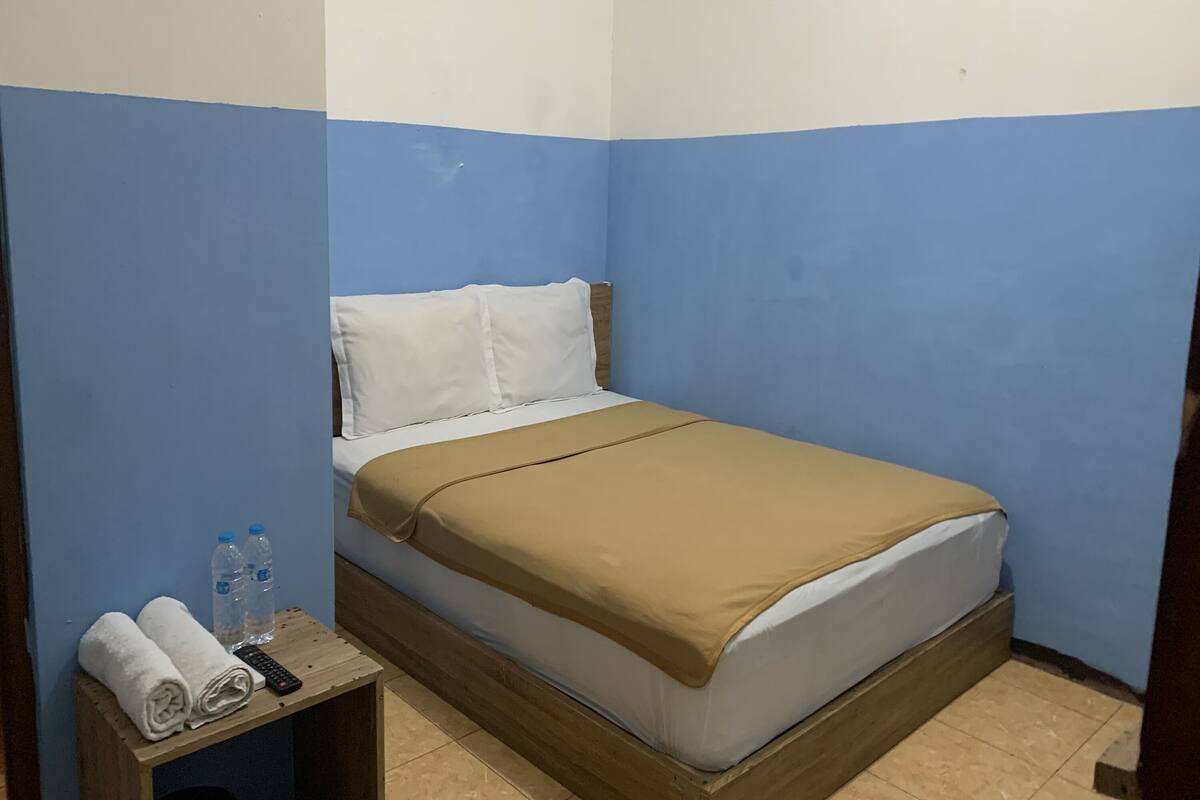 A single bed with a brown blanket, a wooden headboard, and a small wooden nightstand with a towel, two bottles, and a remote control.
