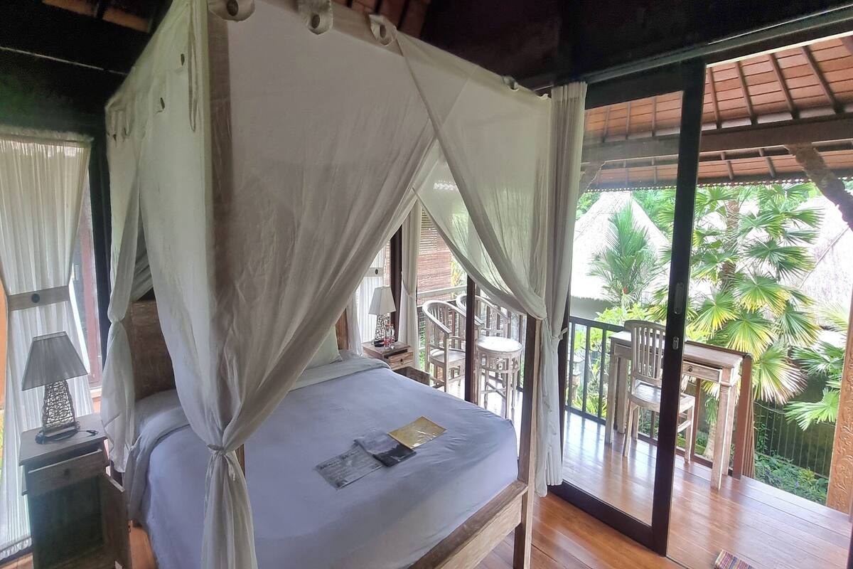 A bed with a canopy, a nightstand with a lamp, and a view of the outdoor balcony with chairs and palm trees.