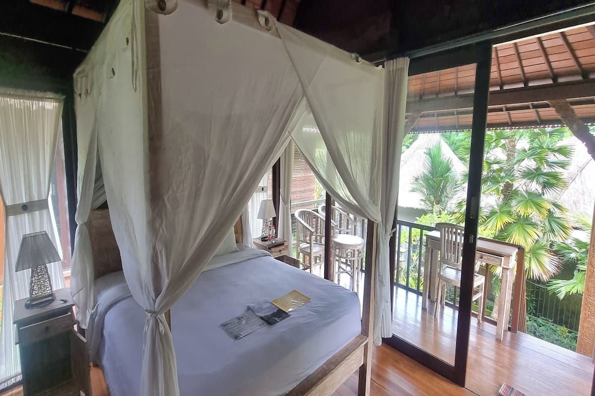 A bed with a canopy, a nightstand with a lamp, and a view of the outdoor balcony with chairs and palm trees.