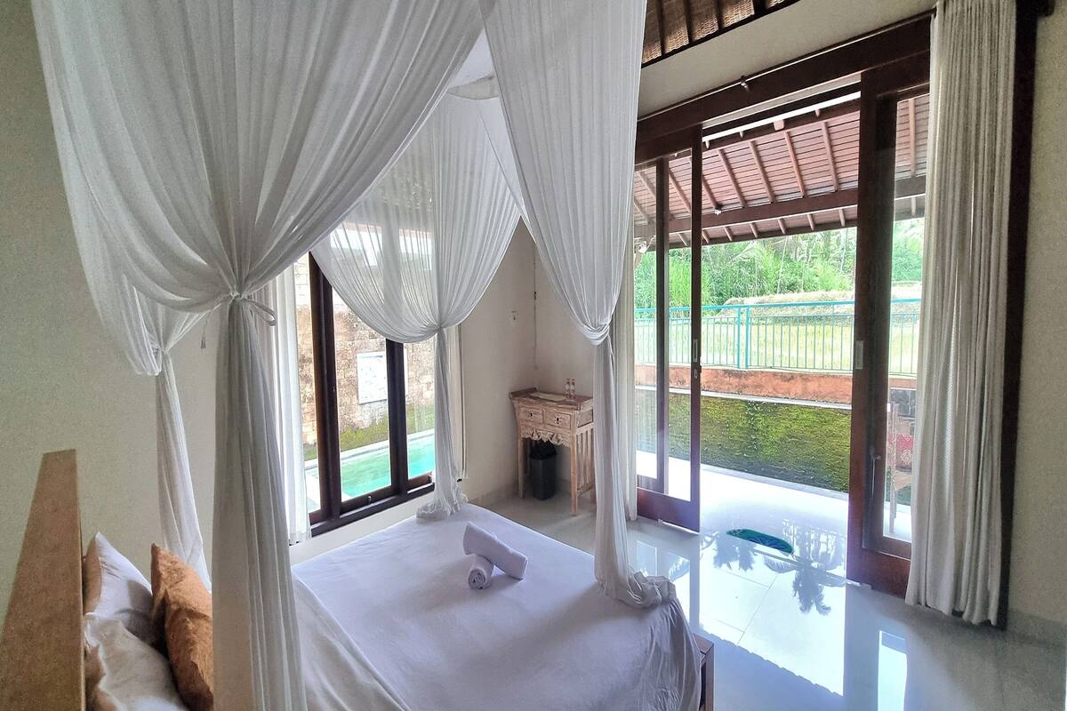 A bedroom with a bed, curtains, a window, and a view of a pool and greenery.