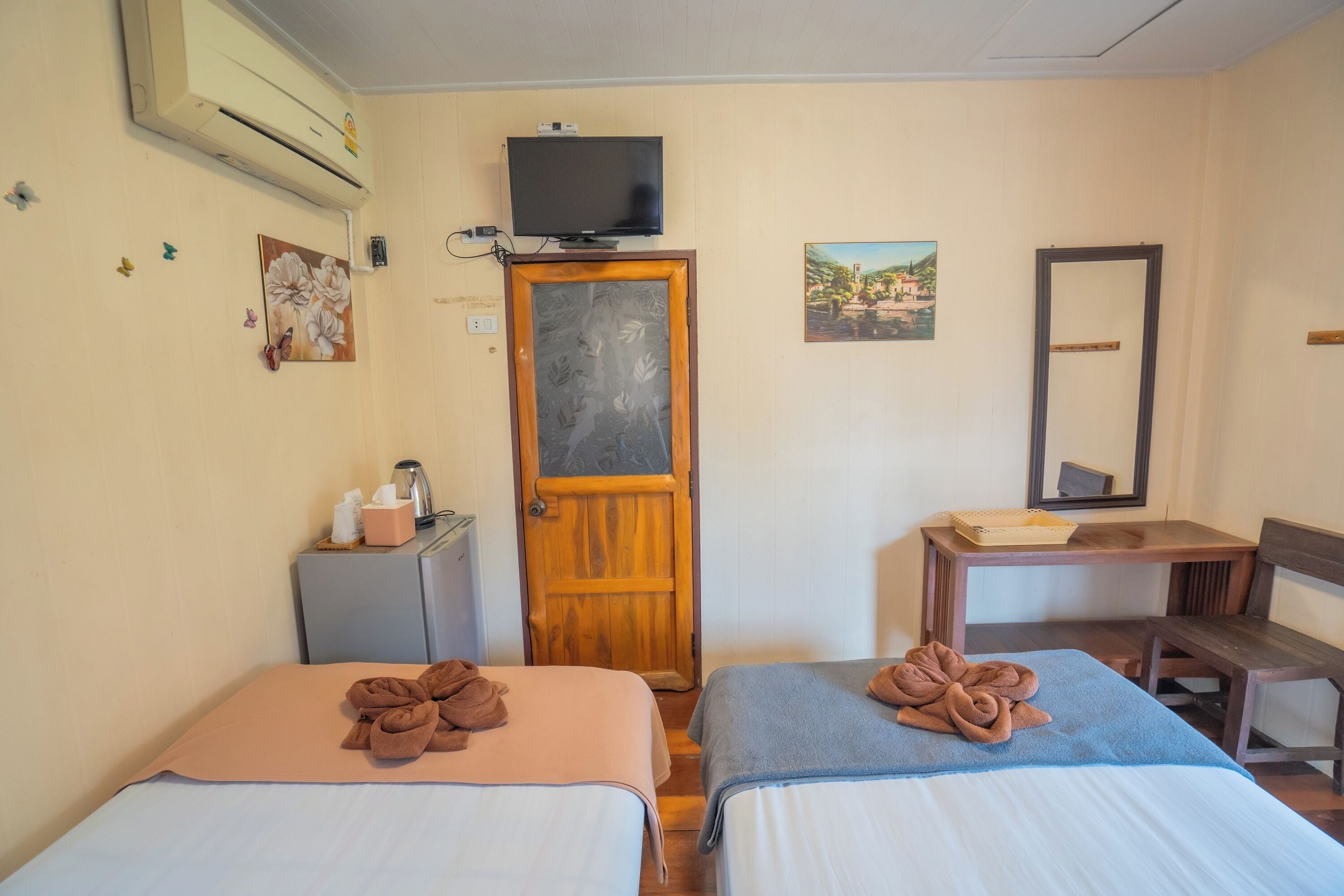 Quarto de hotel com duas camas, uma escrivaninha, uma cadeira, uma televisão, um guarda-roupa e uma porta.