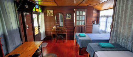 Una habitación con paredes de madera, una mesa de madera, una silla, una cama, una mesita con una lámpara y una ventana con cortinas.