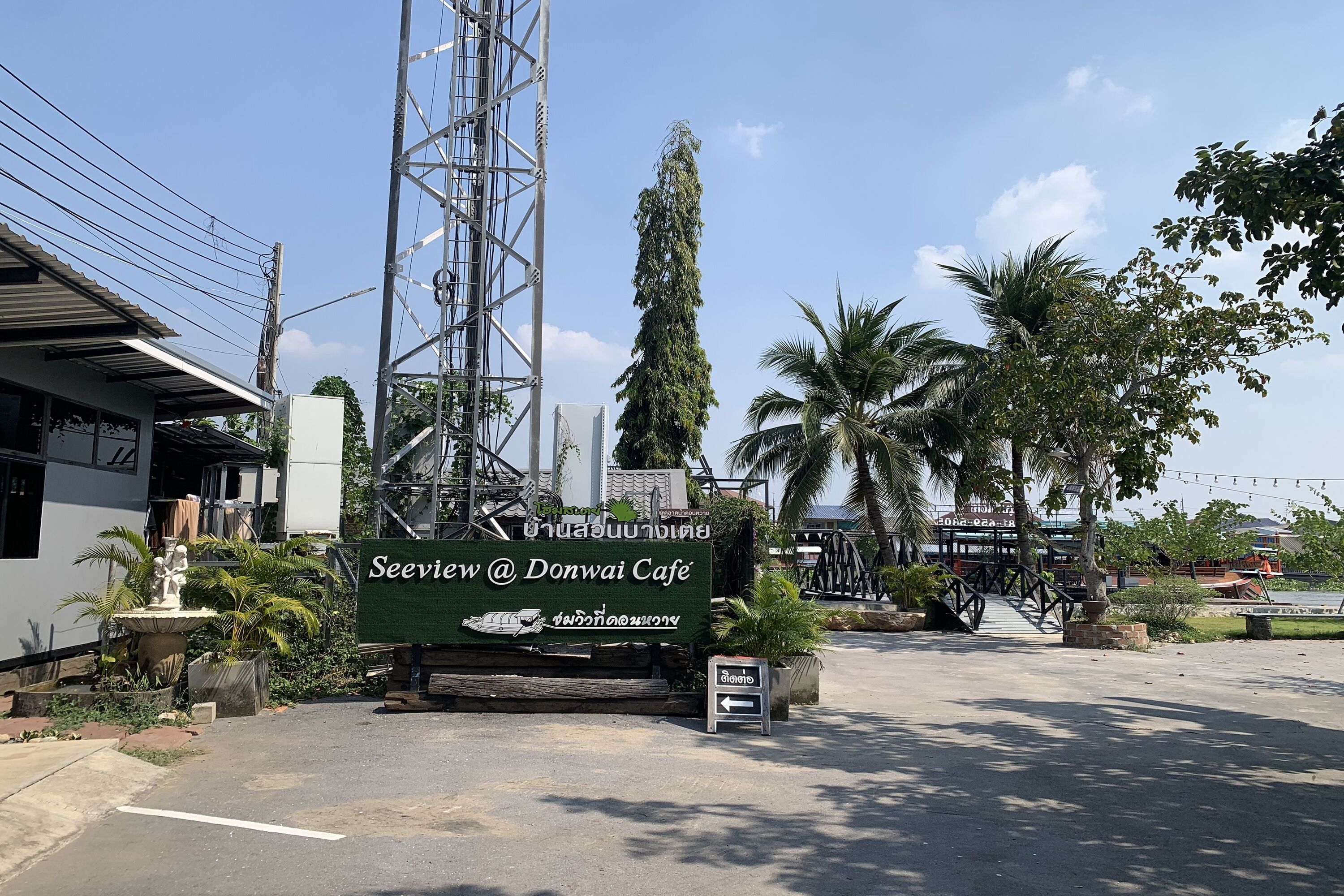 A sign for Seewiew @ Donwai Cafe with a view of the surrounding area including a palm tree, a statue, and a building with a metal roof.