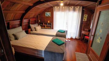 A cozy attic bedroom with two beds, wooden beams, a small desk, and a window with curtains.