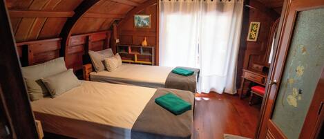 A cozy attic bedroom with two beds, wooden beams, a small desk, and a window with curtains.