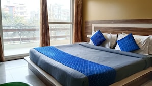 A hotel room with a large bed, two pillows, a window with curtains, and a view of buildings outside. - The Indian Hotel & Kitchen (Gurugram)