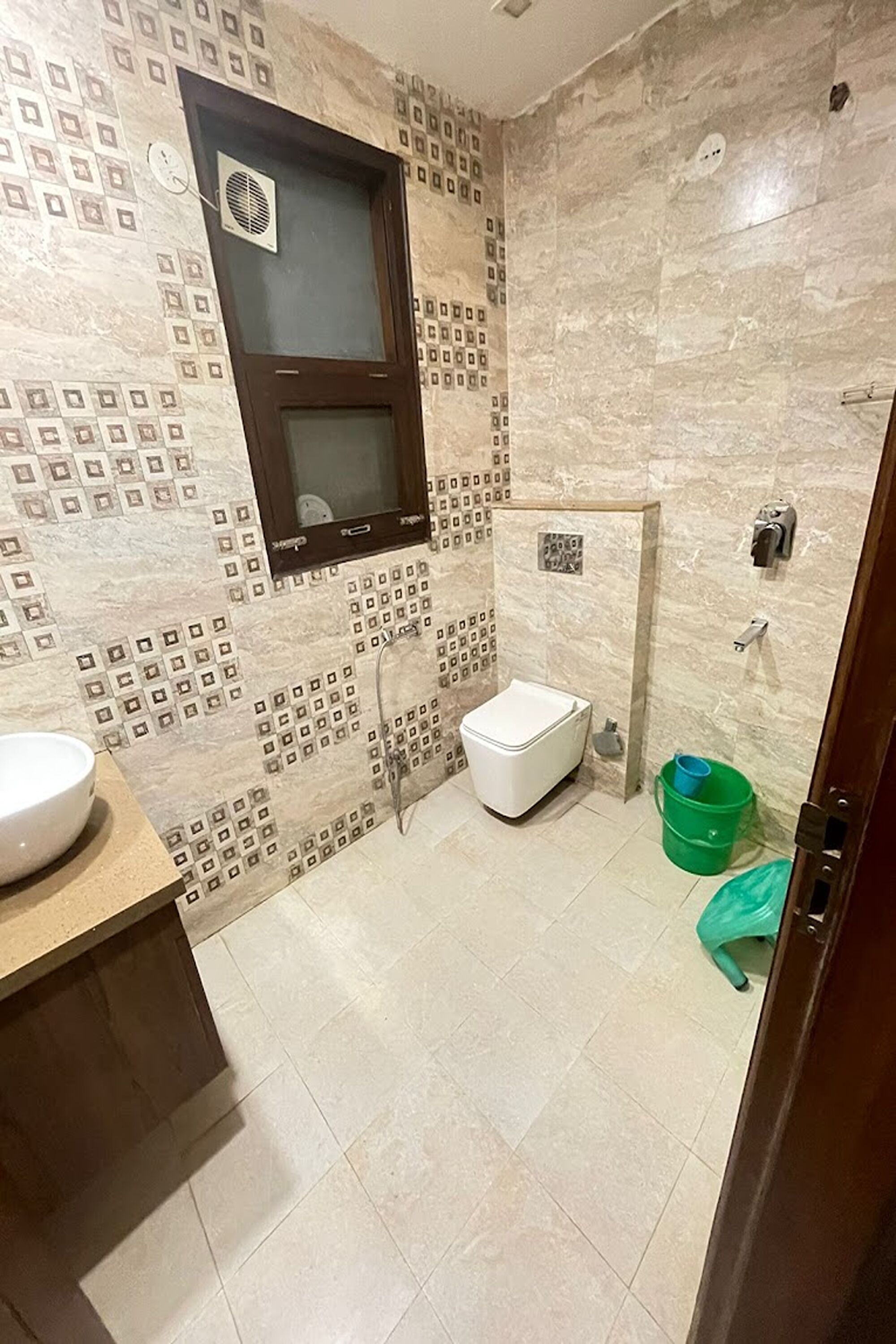 Un baño con baldosas estampadas, un inodoro blanco, un cubo verde y una ventana con ventilador.