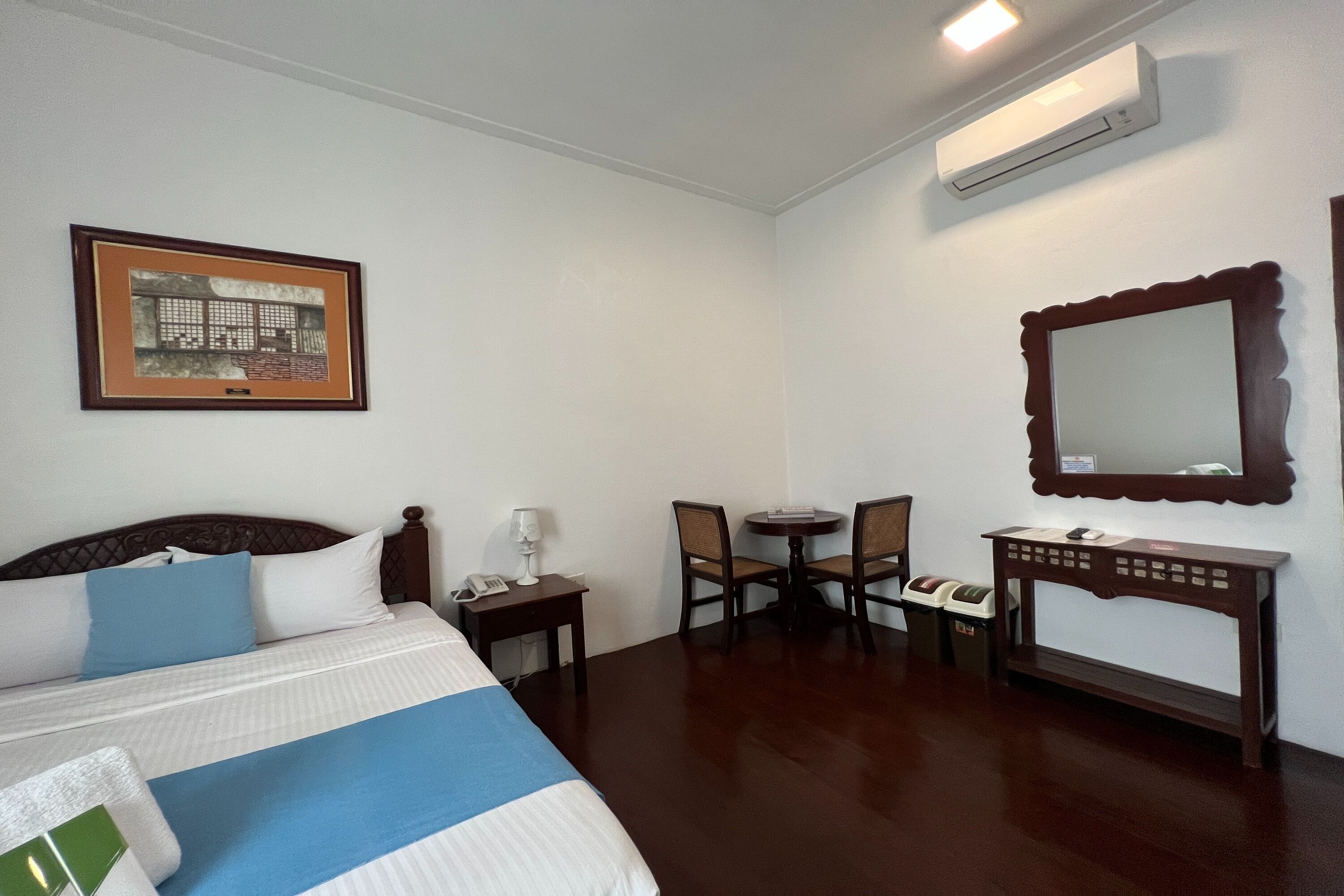 A hotel room with a bed, a desk, a chair, a small table, a mirror, and an air conditioning unit.