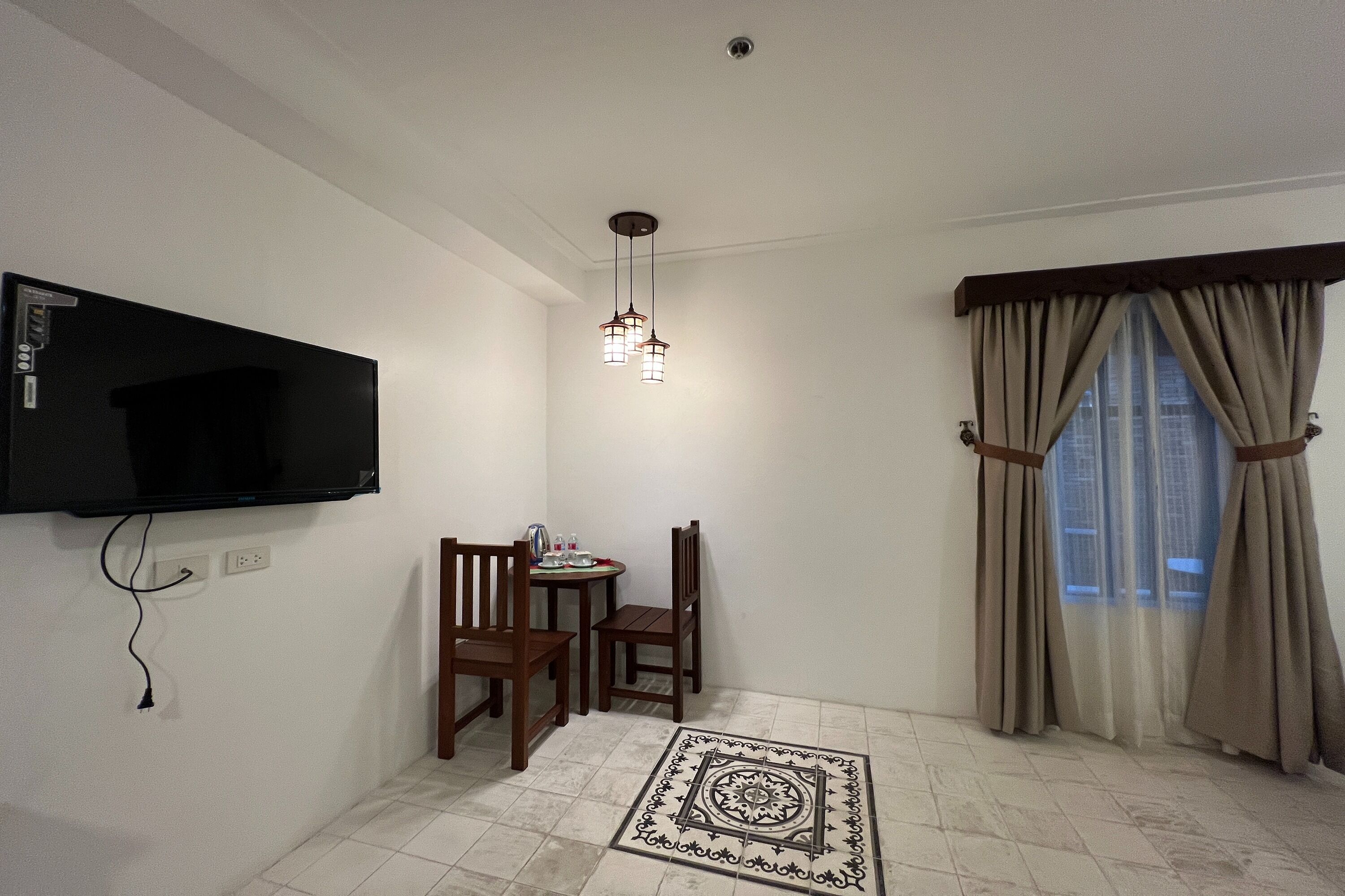 A room with a flat-screen TV mounted on the wall, a dining table with chairs, a patterned rug, and a window with curtains.