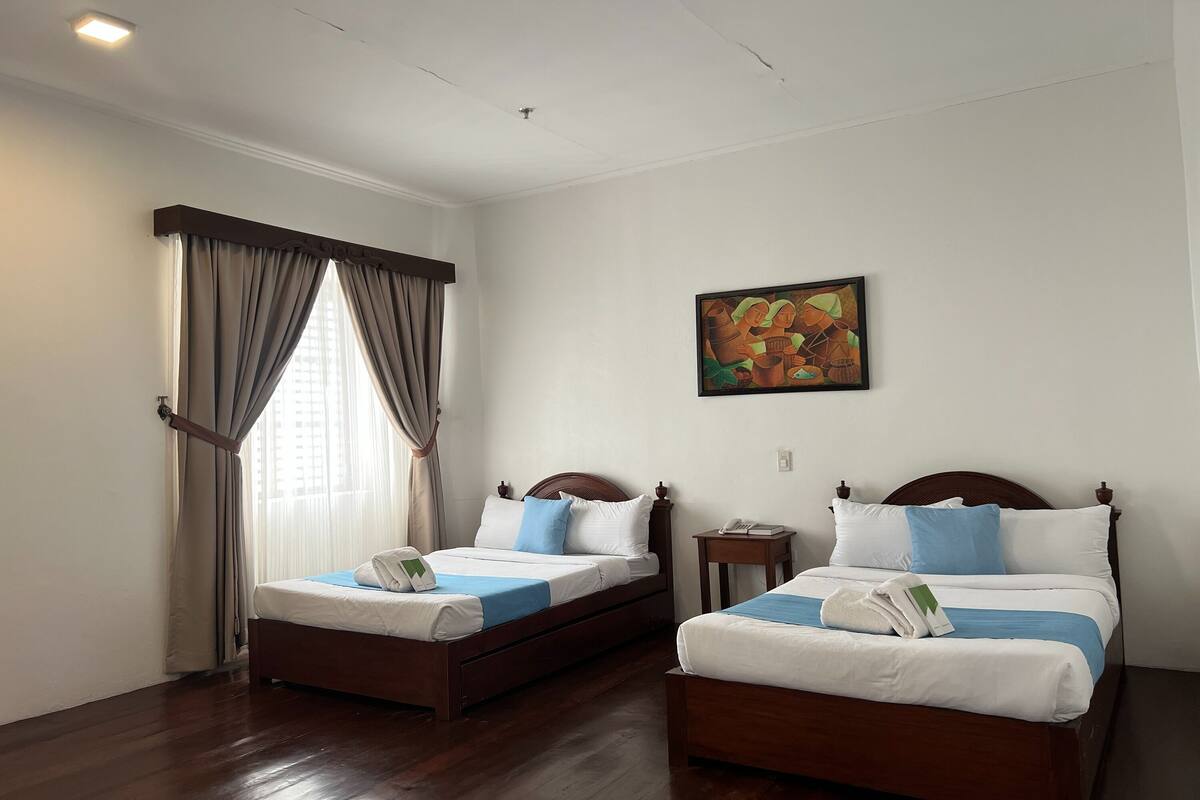 A hotel room with two wooden beds, a window with curtains, a painting on the wall, and a small bedside table with a phone.