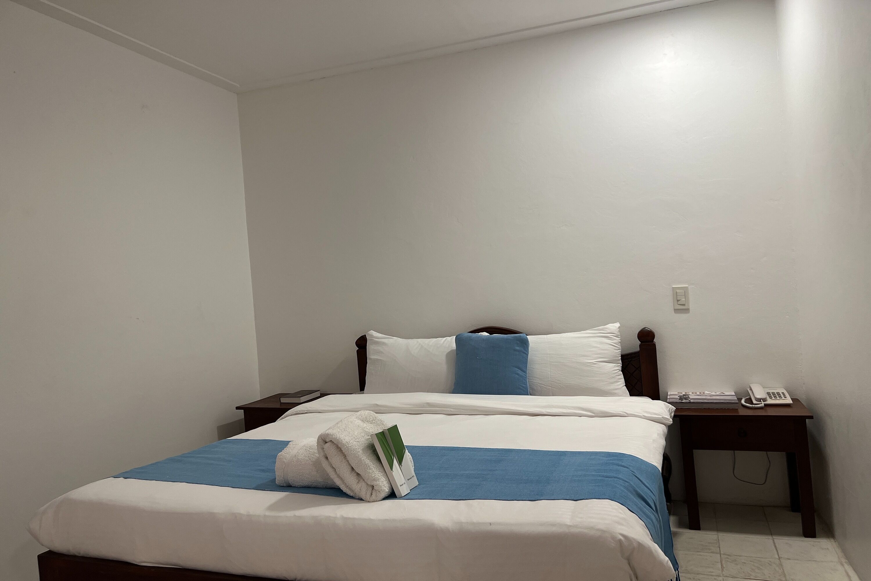 A neatly made bed with a blue and white bedspread, a white towel, and a small card on the side. There is a bedside table with a phone and a book.