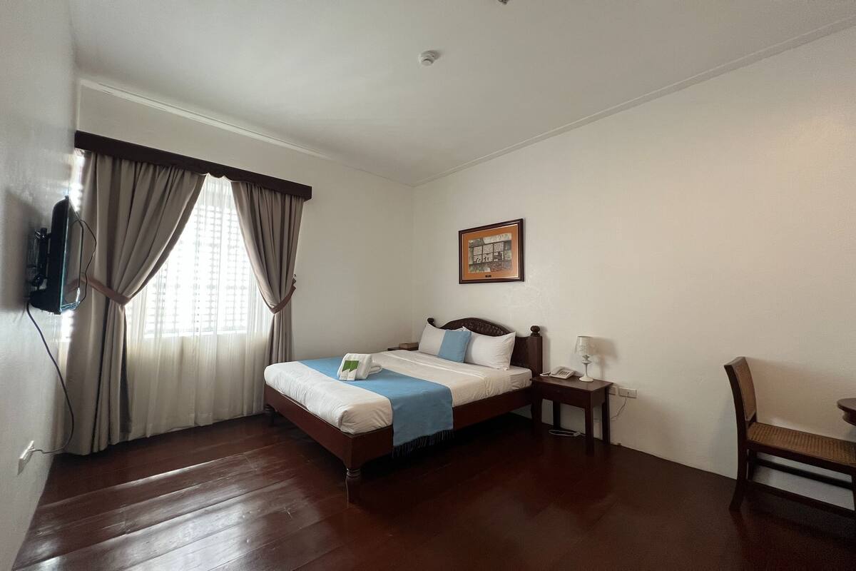 A hotel room with a wooden floor, a bed with a blue and white bedspread, a nightstand with a lamp, a chair, and a framed picture on the wall.