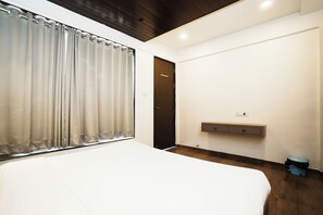 A bedroom with a bed, a window with curtains, a door, a small bedside table, and a trash bin.