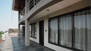 Uma área externa coberta com piso de azulejos, grandes janelas com cortinas e uma varanda com grades.
