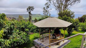 Um gazebo com piso de azulejos e grade de metal, cercado por vegetação exuberante e vista para colinas ao longe.