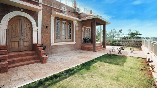 A two-story building with a covered patio, a large wooden door, and a small garden area with outdoor furniture. - Clifton Villa (Mahabaleshwar)