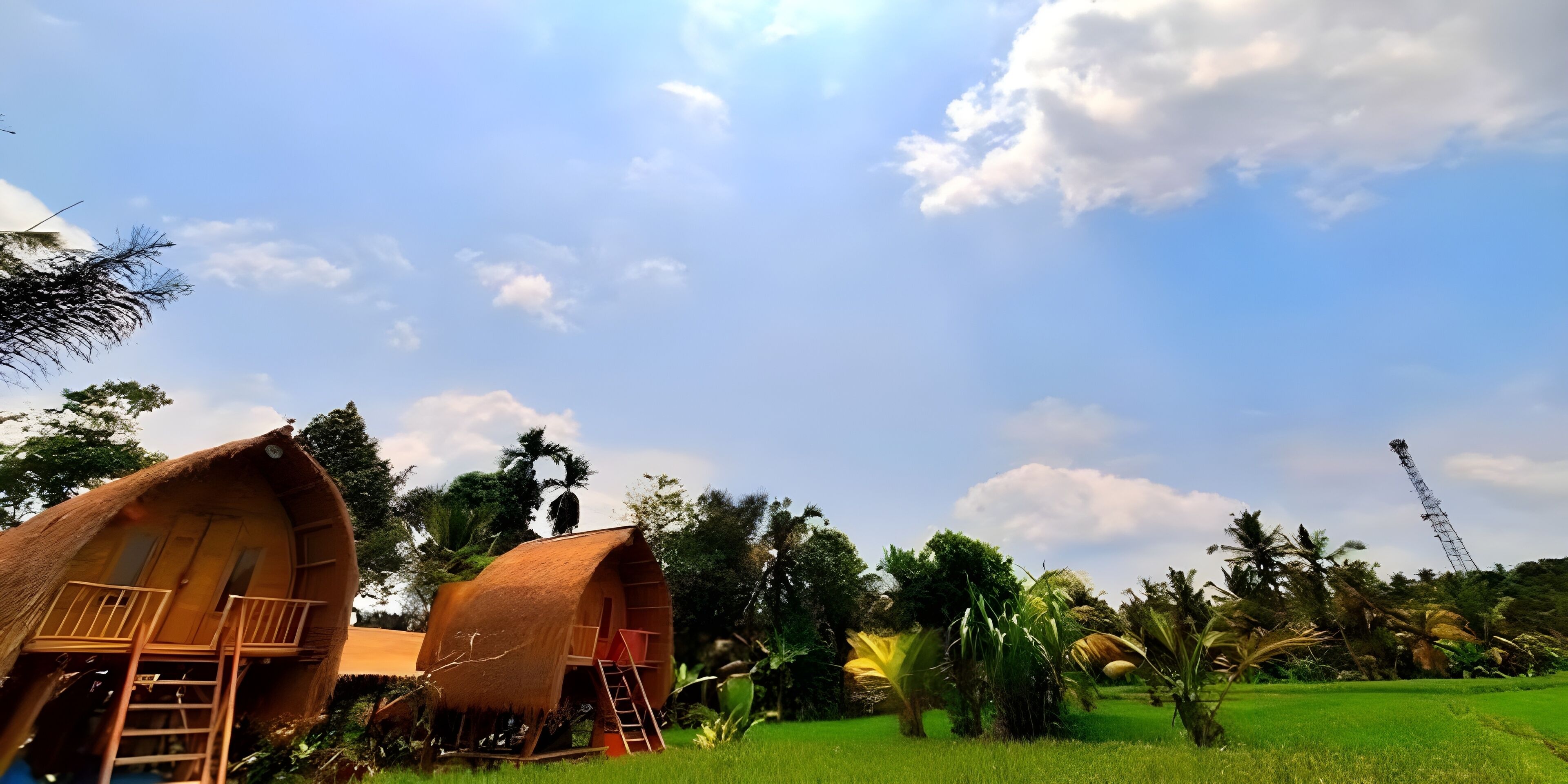 A thatched-roof bungalow with a balcony and a ladder, surrounded by lush greenery and a clear sky.