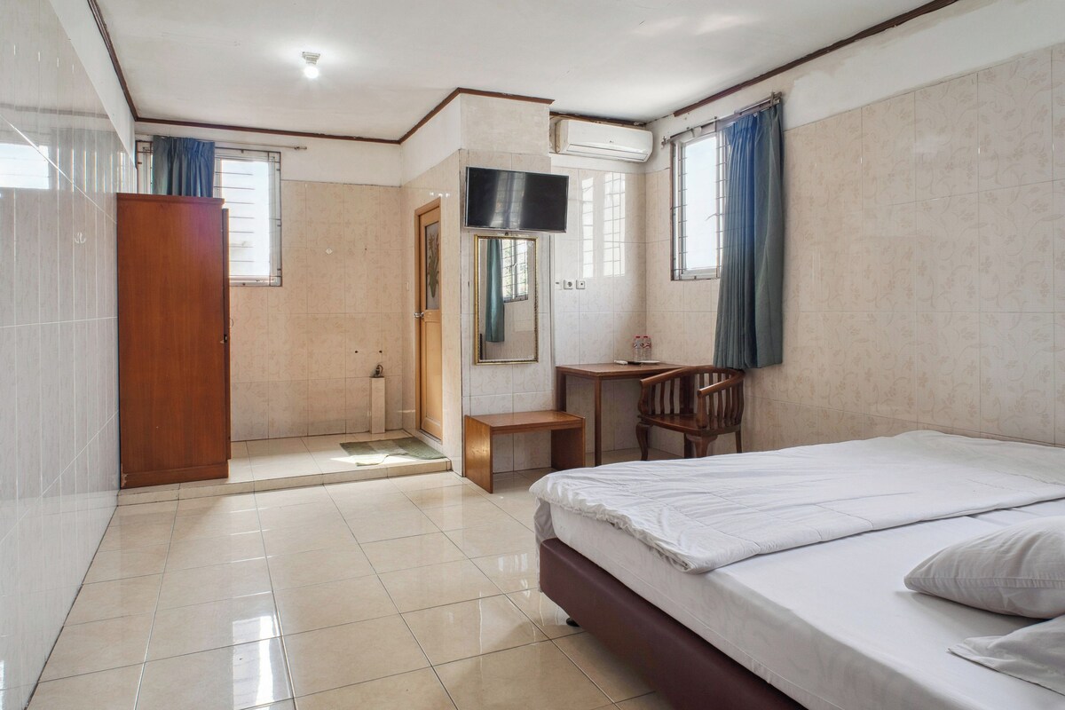 Quarto de hotel com cama, escrivaninha, cadeira, mesinha de centro, armário, chuveiro e televisão.