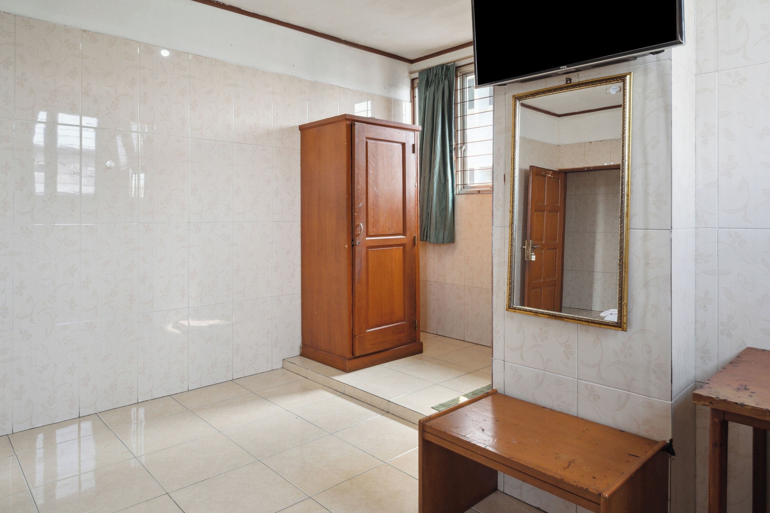 Een kamer met een houten garderobe, een bankje, een spiegel met gouden lijst en een aan de muur bevestigde televisie.