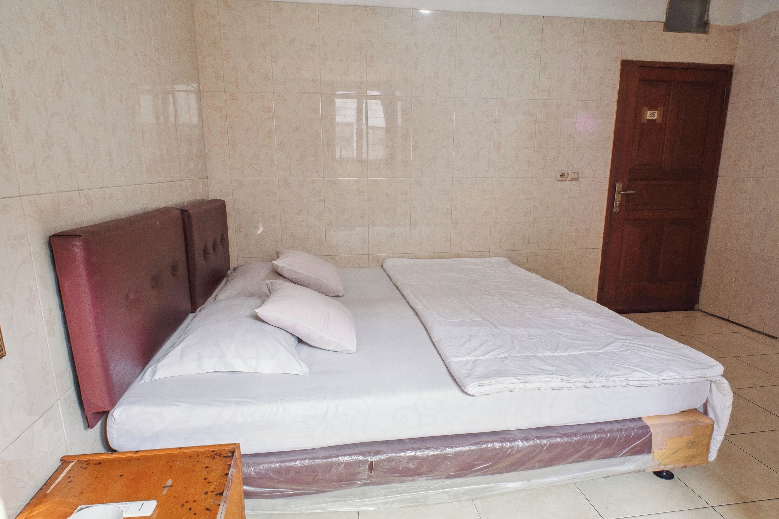 Cama bem arrumada com cabeceira, mesa de cabeceira de madeira e porta em um quarto com paredes revestidas de azulejos.