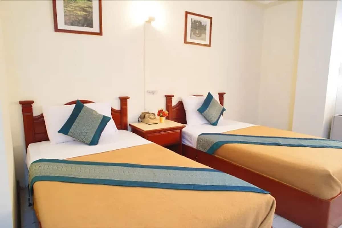Une chambre d’hôtel avec deux lits, chacun doté d’une tête de lit en bois et d’une couvre-lit bleue et beige. Une table de chevet avec une lampe et un téléphone se trouve entre les deux lits.