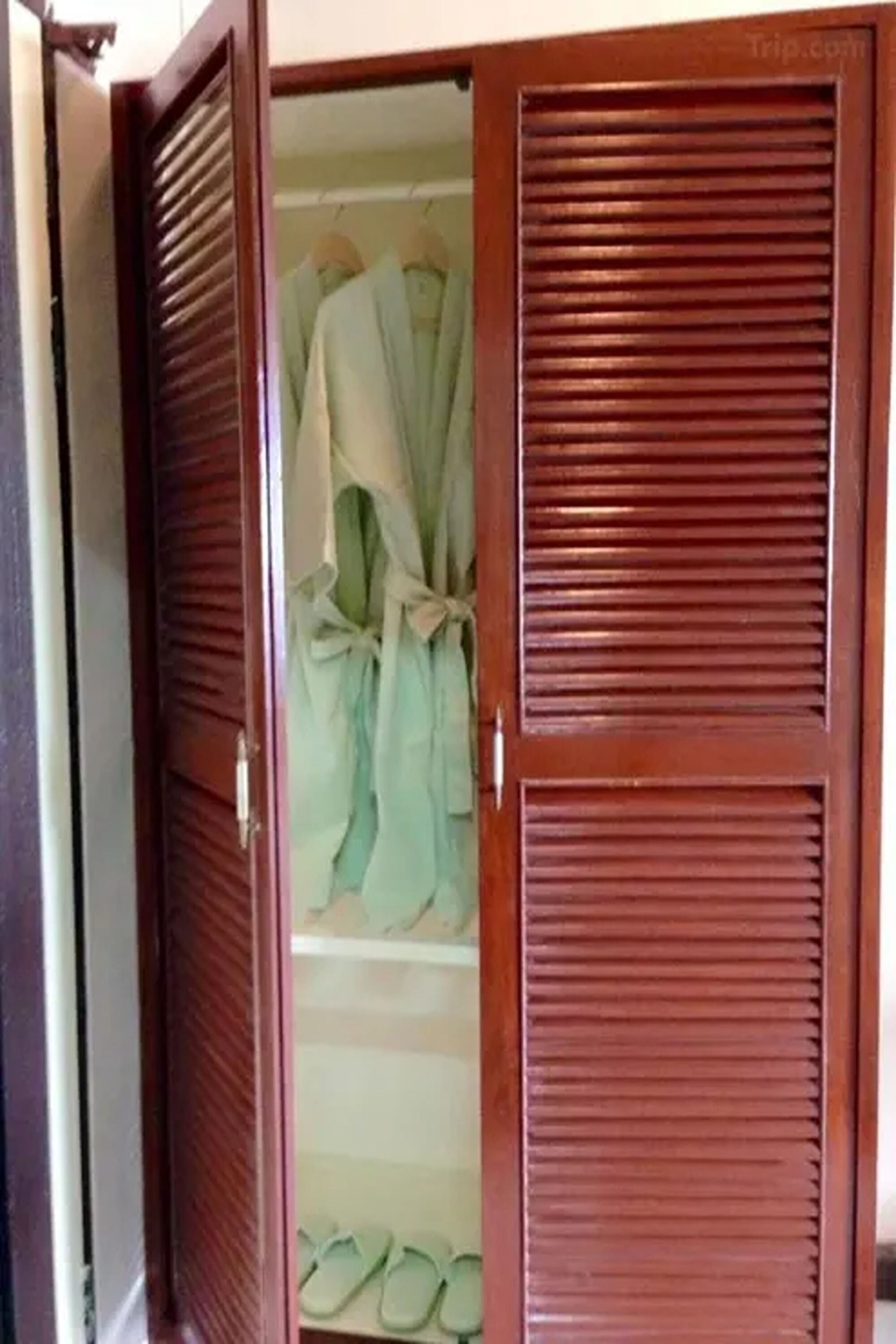 Ein Holzschrank mit horizontalen Lamellen, gefüllt mit ordentlich zusammengefalteter Kleidung und einem Paar grüner Hausschuhe.