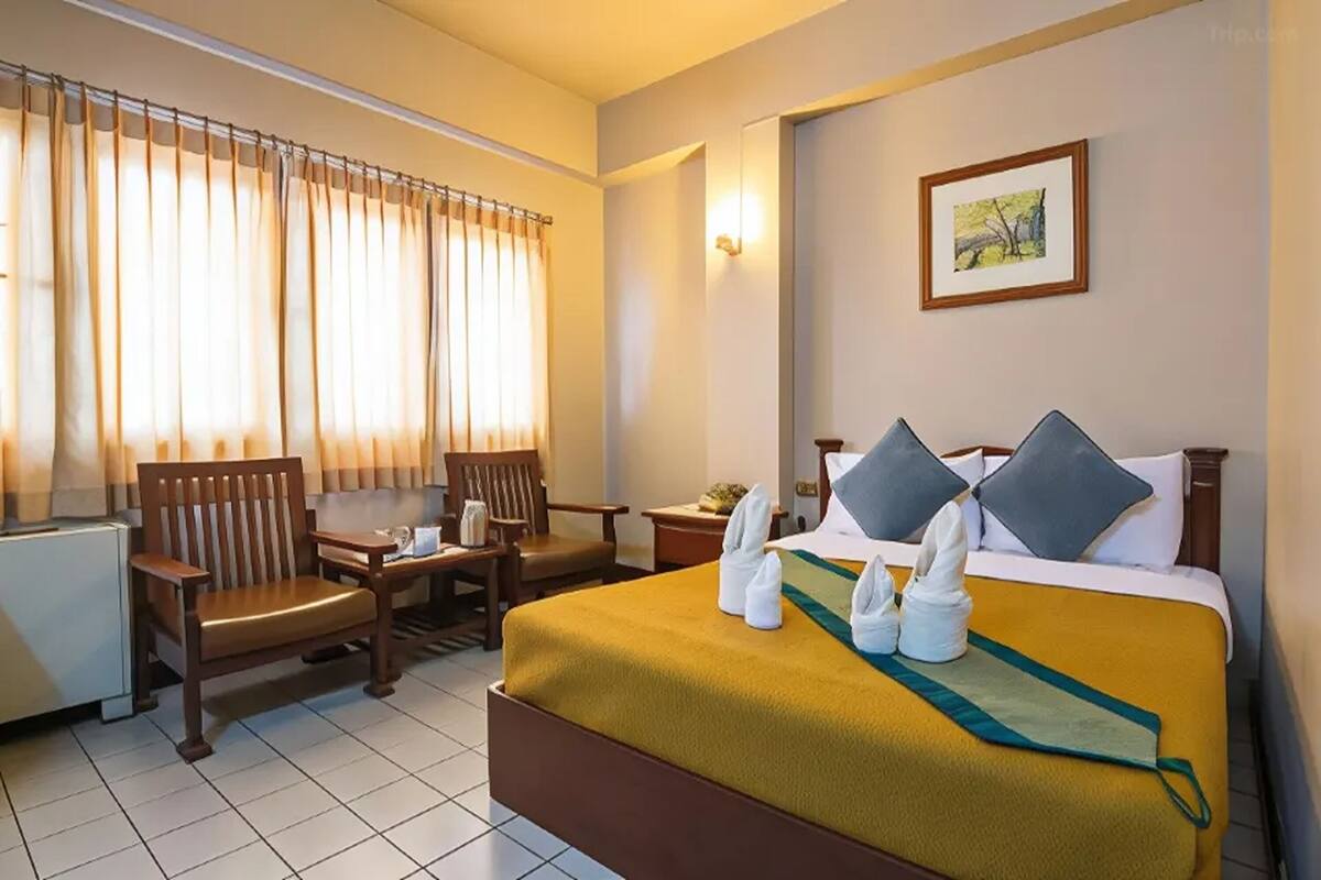 Une chambre d’hôtel avec un lit, deux chaises, une table, un tableau au mur et une fenêtre avec des rideaux.