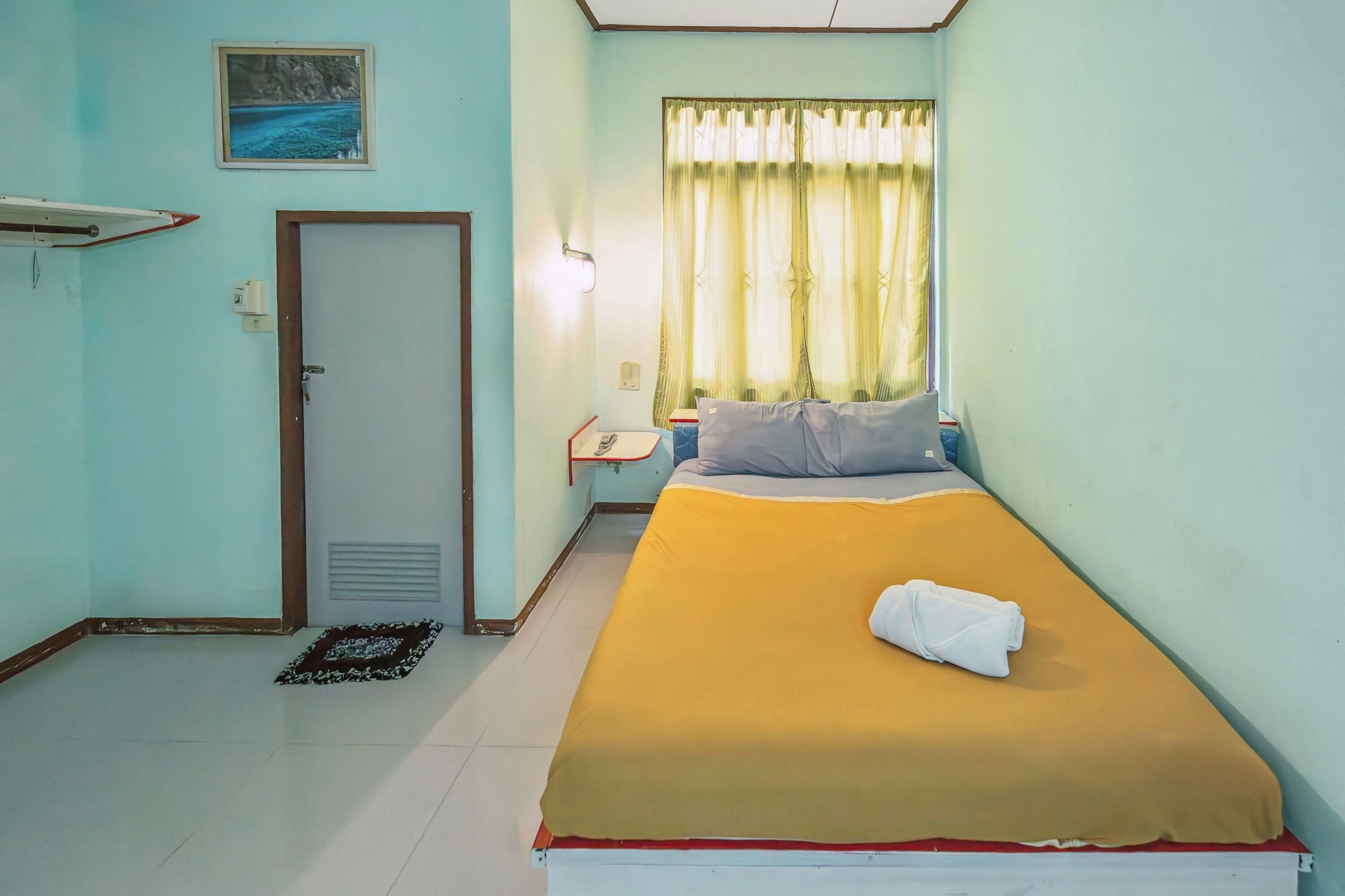 A room with a bed, a nightstand, a window with curtains, a door, and a wall-mounted shelf with a picture.