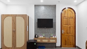 A room with a wooden wardrobe, a flat-screen TV mounted on a grey wall, a wooden door, and a small wooden table with a kettle, cups, and bottles. - Mai Hotel (Ho Chi Minh City)