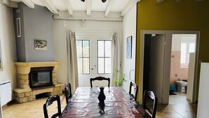 Une salle à manger avec une longue table, des chaises, une cheminée et une vue sur l’extérieur grâce à de grandes fenêtres.