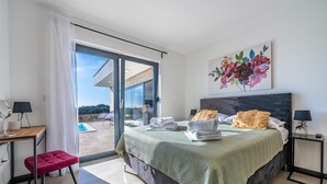 Een moderne slaapkamer met een groot bed, een bureau en een stoel. Er hangt een schilderij aan de muur en er is uitzicht op de omgeving door een schuifdeur van glas.