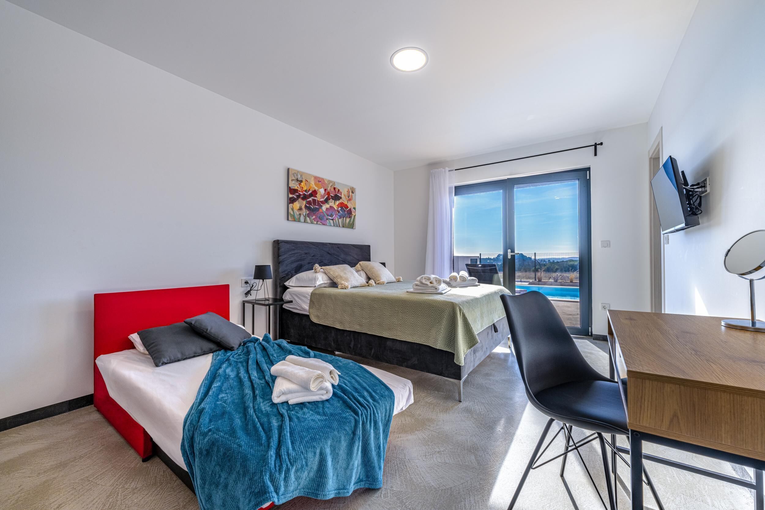 Une chambre d’hôtel moderne avec un lit rouge, un deuxième lit recouvert d’une courtepointe verte, un bureau en bois et une grande fenêtre donnant sur la ville.